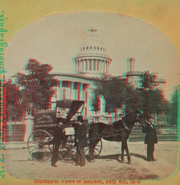 Two men stand by a wagon on which is written "A.L. Dahl Landscape Photographer." The photographer stands on the left side of the view. The flag on the side of Dahl's wagon is a souvenir of the Philadelphia Centennial International Exhibition of 1876. A wrought iron fence surrounds the Capitol Park. This Capitol building, viewed from West Washington Avenue, was erected in 1857 and replaced in 1913. Caption on stereograph reads: "Centennial Views of Madison, July 4, 1876."
