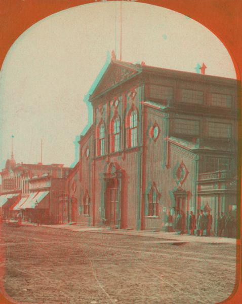 Stereograph. This building, which was destroyed by fire in 1866, was located in the commercial district known as Market Square. The Square was actually a triangular piece of land which extended from East Water Street near Mason to Market Street at Juneau Avenue.  From the middle of the nineteenth century on, it served as a social center for German-American civic life and included shops, beer halls, boardinghouses, and meeting halls. In the image, the large building has wings with roofs that step down two-levels out to each side. A group of people stand on the right.  On the left are more storefronts with awnings.