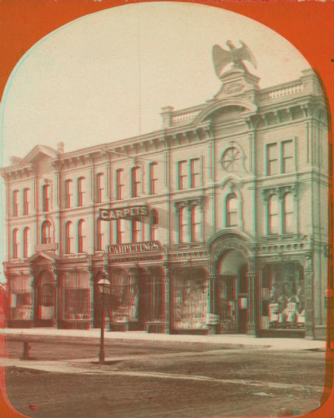 Stereograph; view of store and adjoining buildings, Wisconsin and Milwaukee Streets. A large statue of a bird sits atop the store in the right of the image. Store windows are visible, as well as signs for carpets.