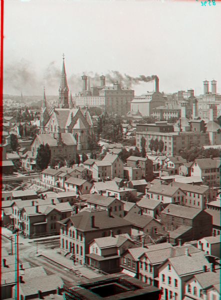 Elevated stereograph looking over the city of Milwaukee. In the background is a church, and factory buildings with smoke flowing out of the chimneys.