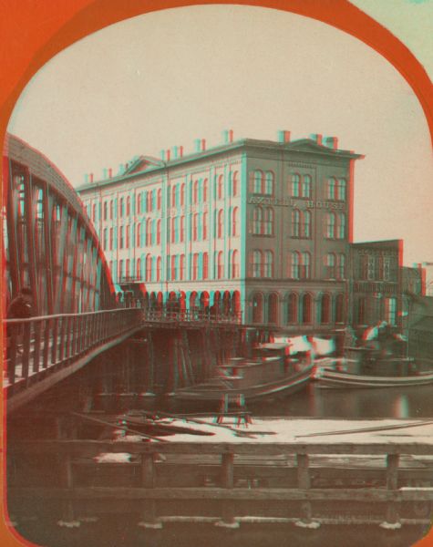 Stereograph of the Axtell House building, with a storefront next to it advertising "Clothing Hats Caps Sailors Outfits".  A bridge is on the left and a man is standing on it. Several boats are on the river, and some snow can be seen on pilings and piers. Located at the corner of Ferry and S. Water Street and seen from E. Water Street.