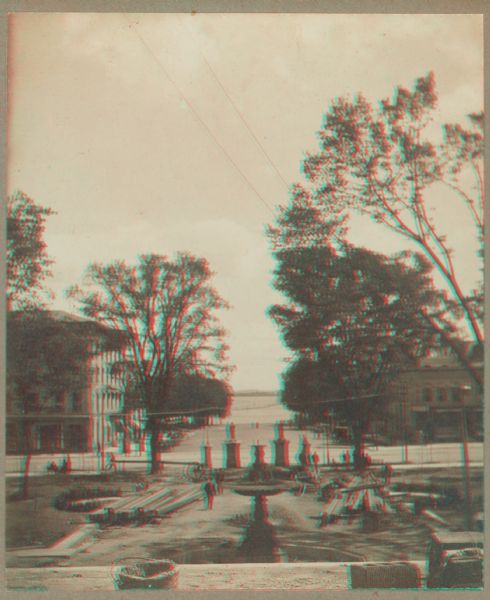 Stereograph view toward Lake Monona from the State Capitol during the construction of the fourth capitol building. The image features the Capitol Square sidewalk (under construction), leading toward a water fountain. Lake Monona is on the horizon.