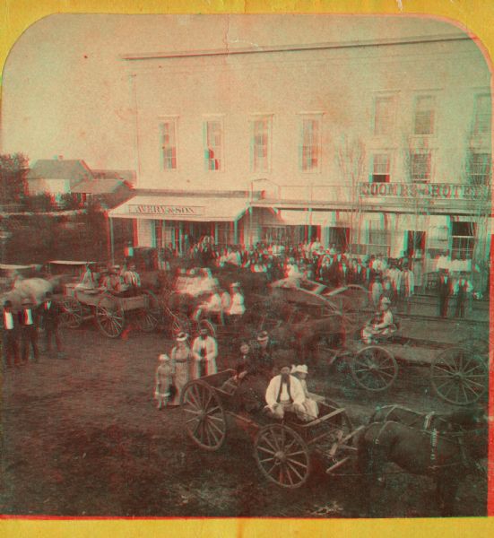 Stereograph of New Richmond(?) during a fourth of July celebration. Identification has not been fully verified, but there was a Coombs Hotel in this town at this time; and other factors also support this identification. Includes horse-drawn carts, crowds of men, women, and children, and two businesses (one with sign reading "Avery & Son" and the other, "Coombs Hotel").