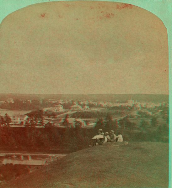Stereograph view of Menomonie. A group of people sit on a hill in the foreground overlooking the town.