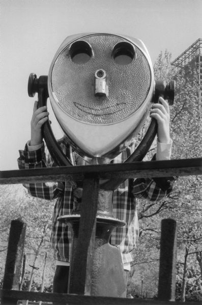 A boy looking through a pay binocular stand in Battery Park.  Graffiti makes the device seem to smile and young trees grow in the background.