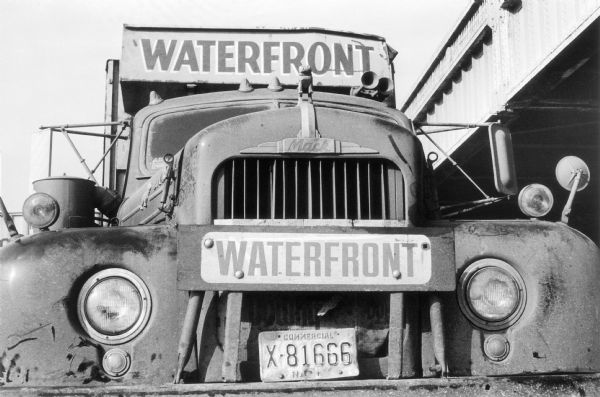 Close-up of the front of a Mack truck, centering on the grille and featuring the Mack bulldog hood ornament. The truck is on West Street parked next to a bridge overpass. The truck has two metal signs that read "Waterfront" and its body sports a layer of grime.