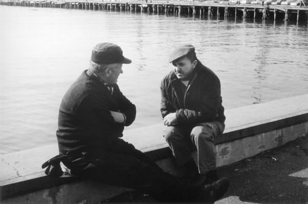 Two men sit in conversation on a low wall beside a watery expanse of the Hudson River.  They are are hunched against the cold and pier pilings form the the top border of the photograph.