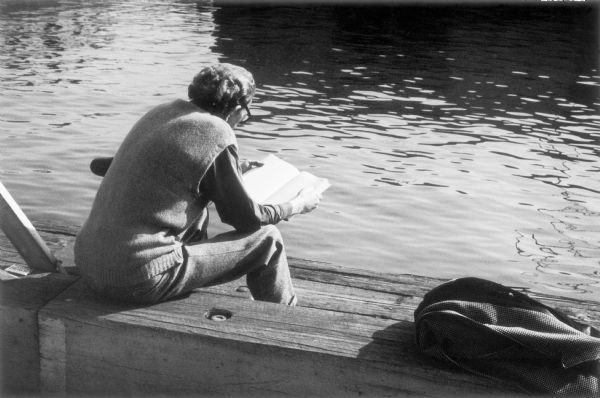 A man in a sweater vest, wool pants and eyeglasses sits in the sun on the pier reading.  Gently rippling water fill most of the photograph while the foreground is composed of the wooden pier and bench.