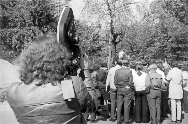 A small crowd gathered around a slightly elevated speaker in Washington Square Park. A videographer recording the event is in the foreground, and park trees bordered by chain-link fence form the background of the scene.