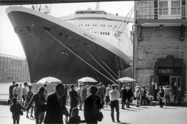 A small crowd mills about a cobblestone plaza near a brick building on the wharf adjacent to the Hudson River. The <i>Queen Elizabeth 2</i> cruise ship is docked and her bow, hull and control room loom over the scene.