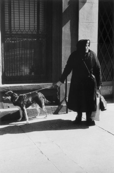 A woman in a thick black coat with a dog on a leash standing on a sidewalk on West 57th Street. The woman is looking into the distance while the dog tugs on the leash in the other direction. The stone building in the background has a barred window and entryway.
