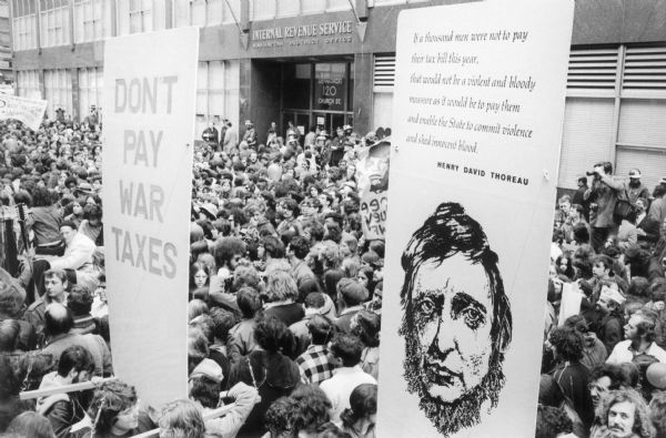 Elevated view of a large crowd gathered in front of the Internal Revenue Service Manhattan District Office to protest war taxes. The protesters carry large signs. One reads, "Don't Pay War Taxes" and the other depicts an anti-war-tax quote from Henry David Thoreau.