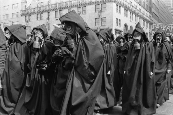 Bread and Puppet Theater members protesting at General Motors Headquarters. A crowd of people wearing gas masks and hooded cloaks reminiscent of the grim reaper walk down the street.  Tall stone buildings and a protest banner are visible in the background.