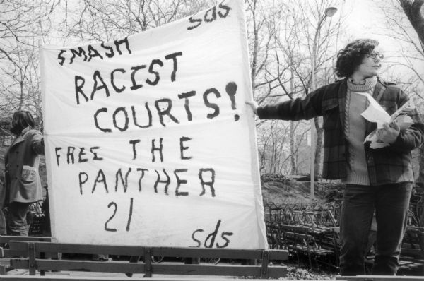 Two protesters hold a banner in Central Park that reads, "Smash Racist Courts! Free the Panther 21, sds." Park benches are stacked on top of each other in the background and the protesters hold leaflets for distribution.