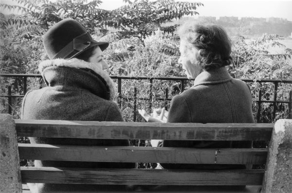Two women sitting on a bench in Riverside Park having a conversation.  The picture is taken from behind with the shoreline far below the women in the background.