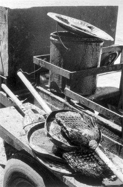 Close-up of tools on the back of a tar wagon on West 181st Street.  Tar soaked brooms glisten in the sun and a pot of tar loosely covered with a hub cap sits nearby.