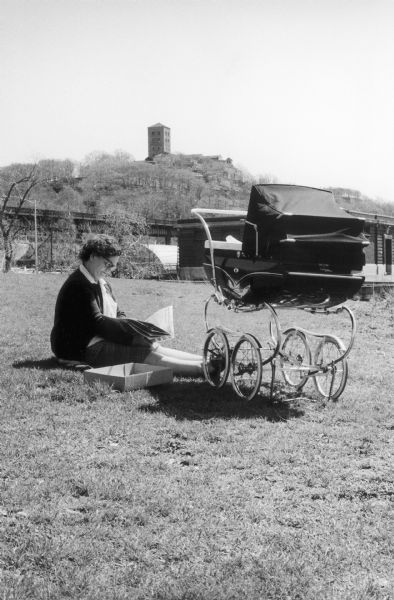 A woman sits on the grass reading next to a baby carriage in Fort Tyron Park. A brush-covered hill surmounted by a square tower rises in the background.