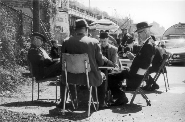 Small groups of men in suits sit around tables in folding chairs playing cards in a sunny parking lot.  A bridge stretches into the distance above the card players.