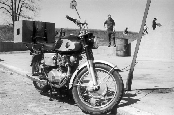 Close-up of a motorcycle with a package lashed to the back is parked at the curb in Inwood Hill Park. Park goers are visible on the grassy hill in the background.