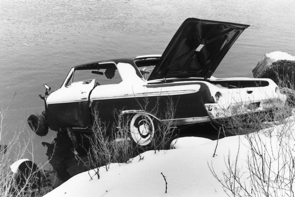 A derelict car overhangs a rocky and snow-bound bank of the Harlem River. Possibly a Ford Galaxie 500 circa 1963 [?]