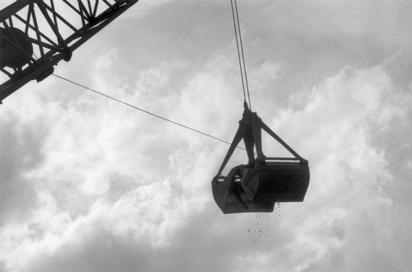 An excavation bucket hanging by cables from a crane at the Harlem River cement works. A cloudy sky forms the background and dirt falls from the bucket.