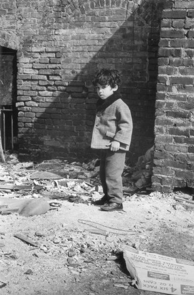 A boy in a short coat stands in front of a brick wall in a debris-strewn alley at East 102nd Street.