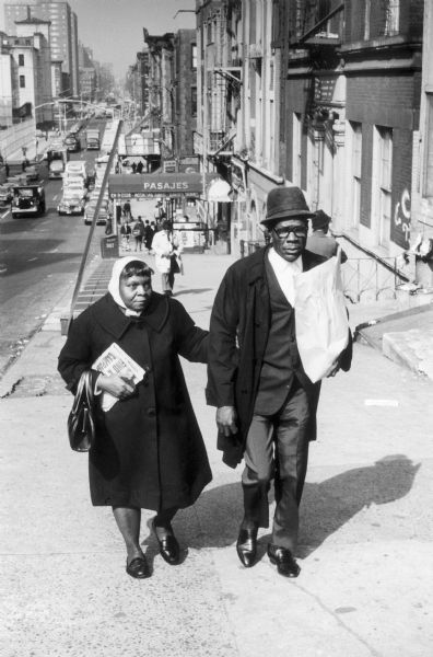 A man and woman walking arm-in-arm up a steep sidewalk. The man is carrying a grocery bag, and the city street lined with buildings stretches off into the distance.