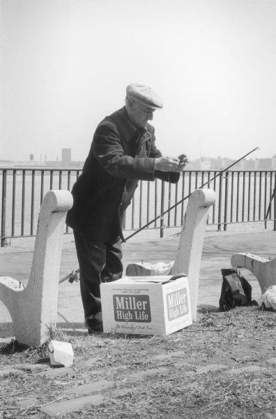 A fisherman baits his hook at a park bench missing its seat.  A metal fence divides the paved shore from the west channel of the East River in the background.