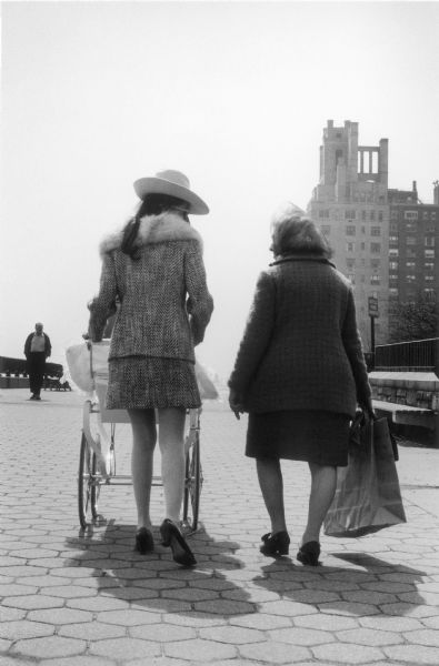 Two women, one holding a bag and one pushing a baby carriage, walk together across the cobbled path of Carl Schurz park.  An approaching pedestrian and the silhouette of a tall building are visible in the distance.