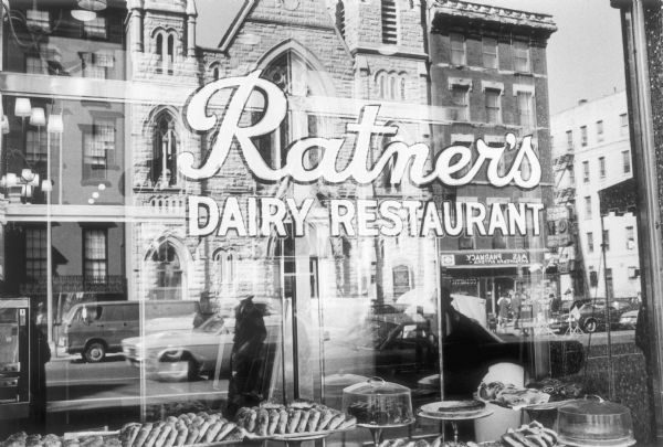 The front window of Ratner's Dairy Restaurant on Second Avenue showing displayed pastries inside and the reflection of buildings and pedestrians on the street.