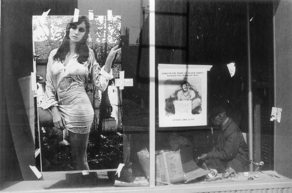 A defunct storefront with a man sitting inside and posters taped in the window.  One poster depicts a woman posing in a wet shirt and the other advertises a meeting.
