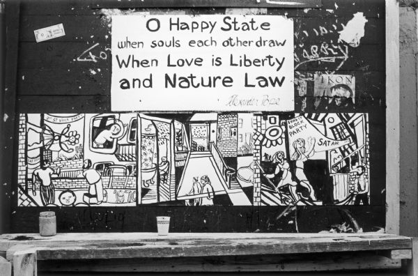 Close-up of a wall with graffiti showing a hand-made poster that reads, "O Happy State when souls each other draw, When Love is Liberty and Nature Law."  Below is a three-panel black and white cartoon strip.