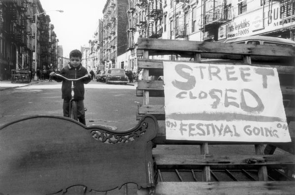 A boy sitting on a bike on the other side of a street barrier.  A pallet and a head board block off the street while a sign announces, "Street Closed--Festival Going On."  In the background a street with parked cars, pedestrians and tall buildings stretches into the distance.