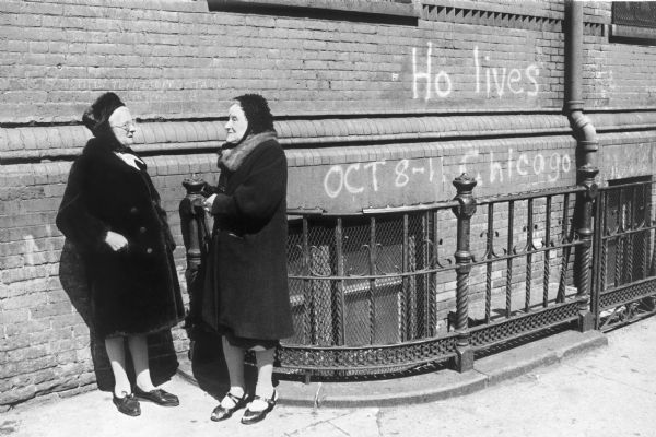 Two women having a conversation standing on a sidewalk next to a brick building and near a decorative metal fence.  Graffiti on the brick wall reads, "Ho lives, Oct. 8-11 Chicago."