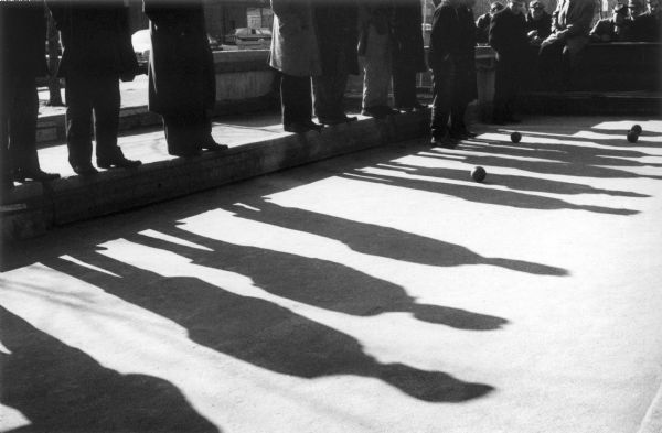 A group of men watch a bocce game on a court on East Houston Street.  The line of men standing on the lip of the court cast elongated silhouettes onto the pavement.