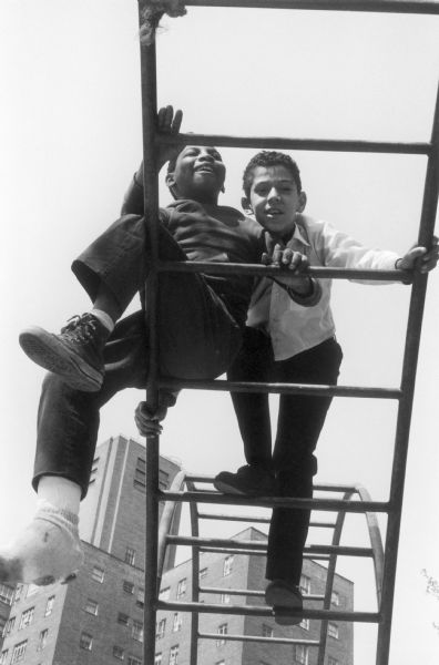 View from below of two boys on a jungle gym.  Some of the brick buildings of the Jacob Riis Houses rise in the background.