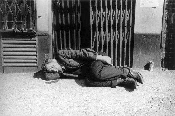 Man sleeping on a sidewalk in front of a retractable metal gate in the Bowery.