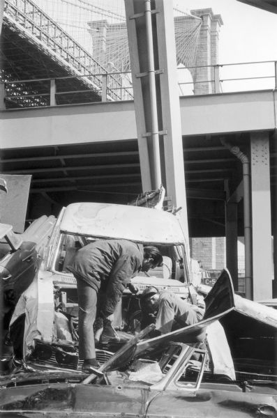 Two men on a derelict car under the Brooklyn Bridge.  They appear to be salvaging parts.  Other wrecked vehicles crowd the area.