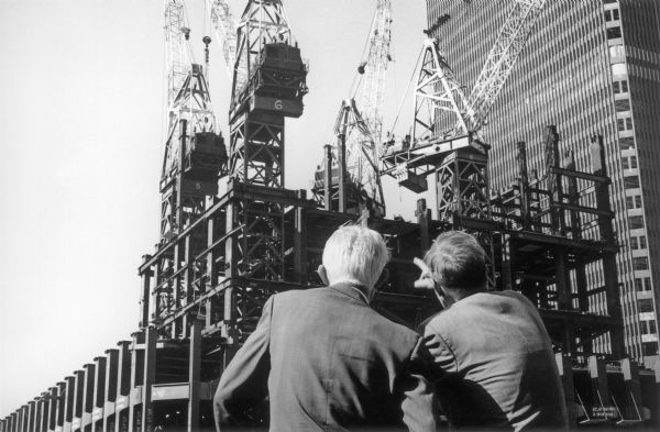 Two men in suits watch four large cranes working to construct the World Trade Center.  The cranes sit atop an extensive metal-beam structure and the men are seen from behind.