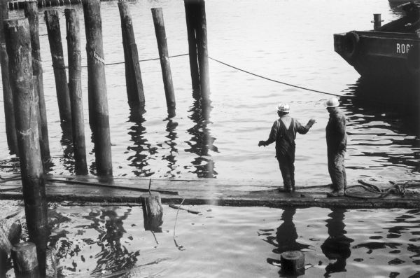 Two workmen in overalls and hardhats stand on a wooden walkway in the East River.  The slick water-level walkway leads into a group of pilings and the back of a metal boat is visible nearby.