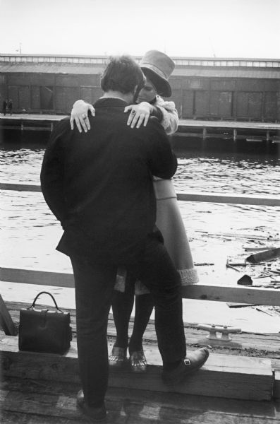 A fashionably dressed young couple stand in an embrace on a pier on the East River.  The river and a waterfront warehouse are visible behind them.