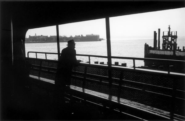 Two passengers stand over a bench and look out of the ferry towards the water.  The developed shoreline and a pier with a seagull perched on top can be seen from the ferry's deck.