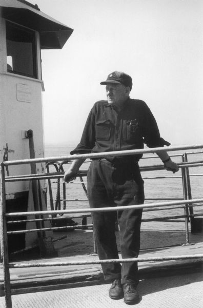 A tugboat captain leans on a handrail and squints into the sun on a tug in the East River.  Part of the wheelhouse is visible behind him.