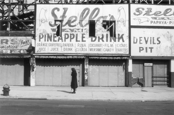 A woman standing in front of closed stalls on Coney Island.  The framework for the Devil's Pit roller coaster rises in the background behind dilapidated signs advertising Stella's famous pineapple drink.