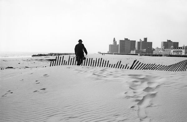 A man walking with a cane in the sand dunes on Coney Island.  A dilapidated fence cuts across the foreground of the beach and a rock jetty juts into the bay in the background.  An amusement park and tall buildings are also visible in the distance.