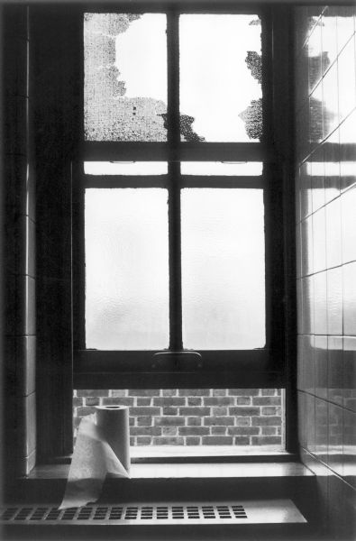 Close-up of a partially open restroom window in the Bellevue Hospital.  A heating vent is attached to the wall in front of the window and roll of paper towels sits on the window sill.  The window panes are frosted and dirty.