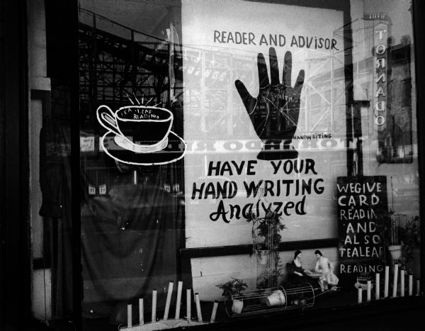 Close-up of a fortune teller's shop front on Coney Island. The multiple signs read, "Reader and Advisor," "Have Your Hand Writing Analyzed," "We Give Card Readings and Tea Leaf Readings." The display features artificial candles and plants as well as figurines seated at a miniature table. The Tornado roller coaster structure reflects on the window's glass.