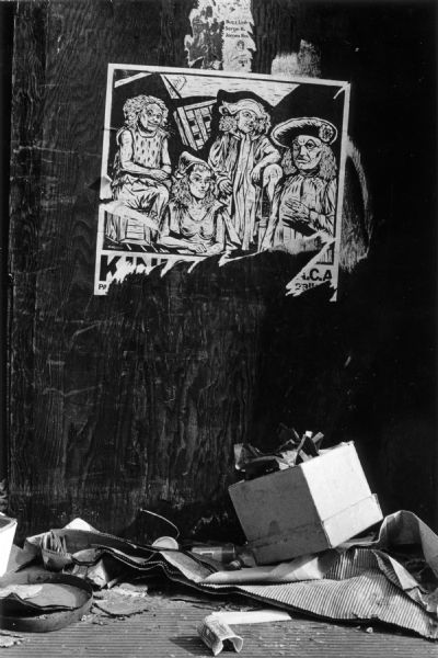 Close-up of a poster attached to a dark wooden surface in the East Village with a layer of trash collected on the ground below.  The poster features a black and white drawing of three men and one woman in historic clothing.  The text portion of the poster is missing.