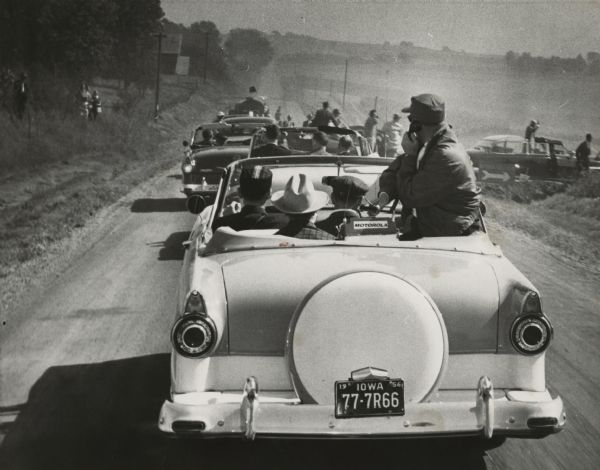 Rear view of a parade of reporters in open cars following President Eisenhower on a rural road in Iowa. The President is standing in an open car at the right.