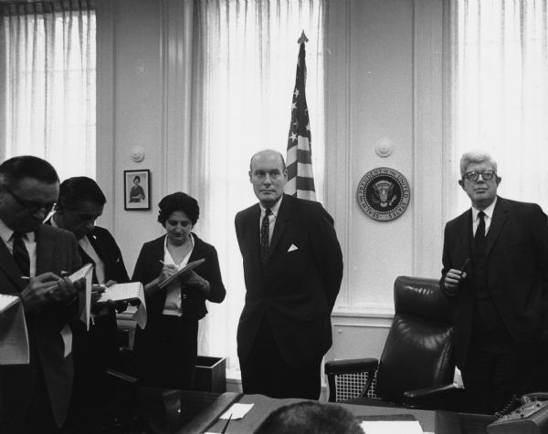 Recently-appointed Attorney General Nicholas Katzenbach answers questions from the press. To his left is George Reedy, press secretary for President Lyndon B. Johnson. Reedy later became a professor in the Journalism School at Marquette University. The journalists include Merriman Smith (second from the left) and Helen Thomas, both reporters for UPI.